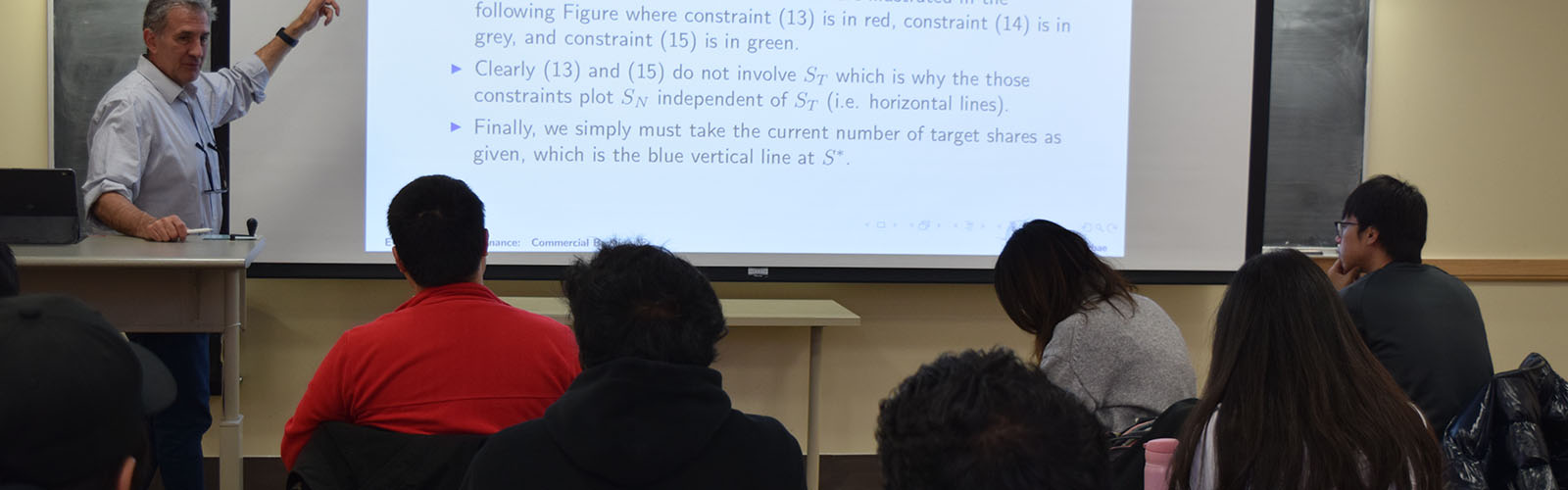 graduate students sit in a classroom, listen and take notes as the professor instructs